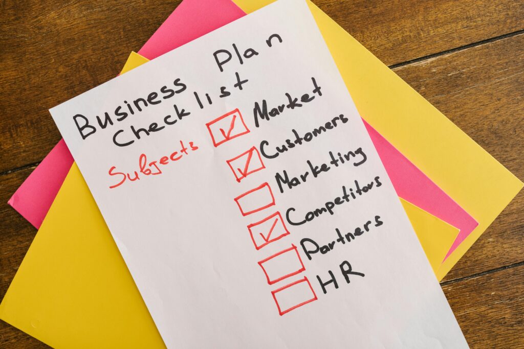 Business plan checklist on colorful sticky notes showing Market, Customers, and Competitors checked off, with Marketing, Partners, and HR remaining