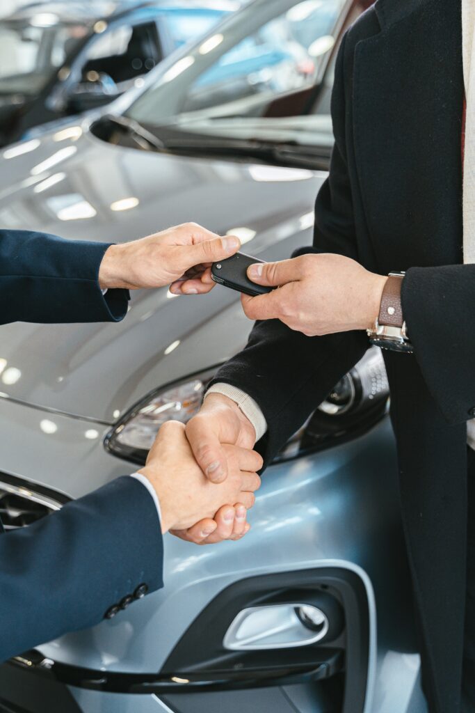Two professionals shaking hands during a car delivery, with one person handing over keys to the new owner, illustrating the intentional moment where advocacy and referrals begin