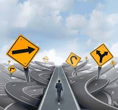 A businessman in a suit walks along a straight road toward a cluster of yellow directional signs pointing in different, tangled directions, with a chaotic maze of overlapping roads swirling in the background beneath a partly cloudy sky — symbolizing confusion, indecision, and the challenge of choosing a clear path forward.